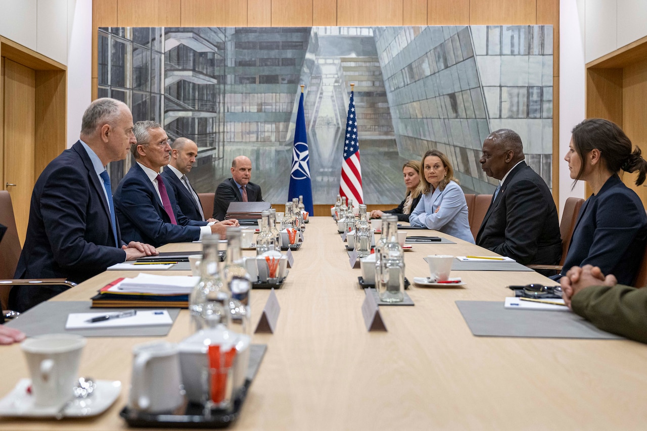 People wearing business attire sit around a conference table with a United States and NATO flag in the background.