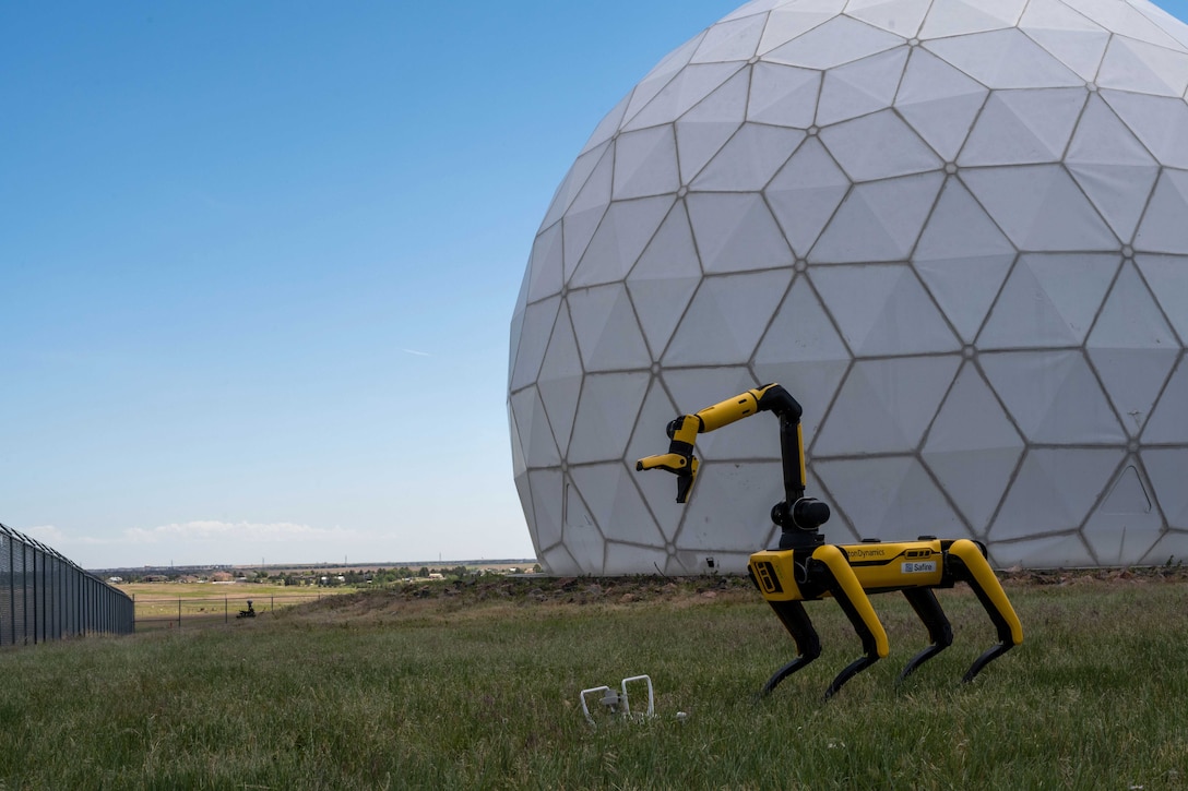 A robot dog extended its neck to pick up a drone in grass with a bubble structure in the background.