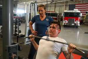 U.S. Air Force Tech. Sgt. Michael Swords, 312th Training Squadron instructor supervisor, is coached by Cheri Cotton, Operational Support Teams strength and conditioning coach, exercise physiologist, and nutritionist, while performing a lat pulldown exercise at the Louis F. Garland Department of Defense Fire Academy, Goodfellow Air Force Base, Texas, March 30, 2024. The base commander strategically decides where to assign OST based on data such as medical needs, medical profiles, low physical training scores and administrative actions that have been taken. (U.S. Air Force photo by Airman 1st Class James Salellas)
