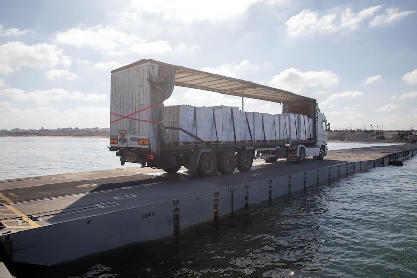 A truck carrying cargo drives on a pier toward land.