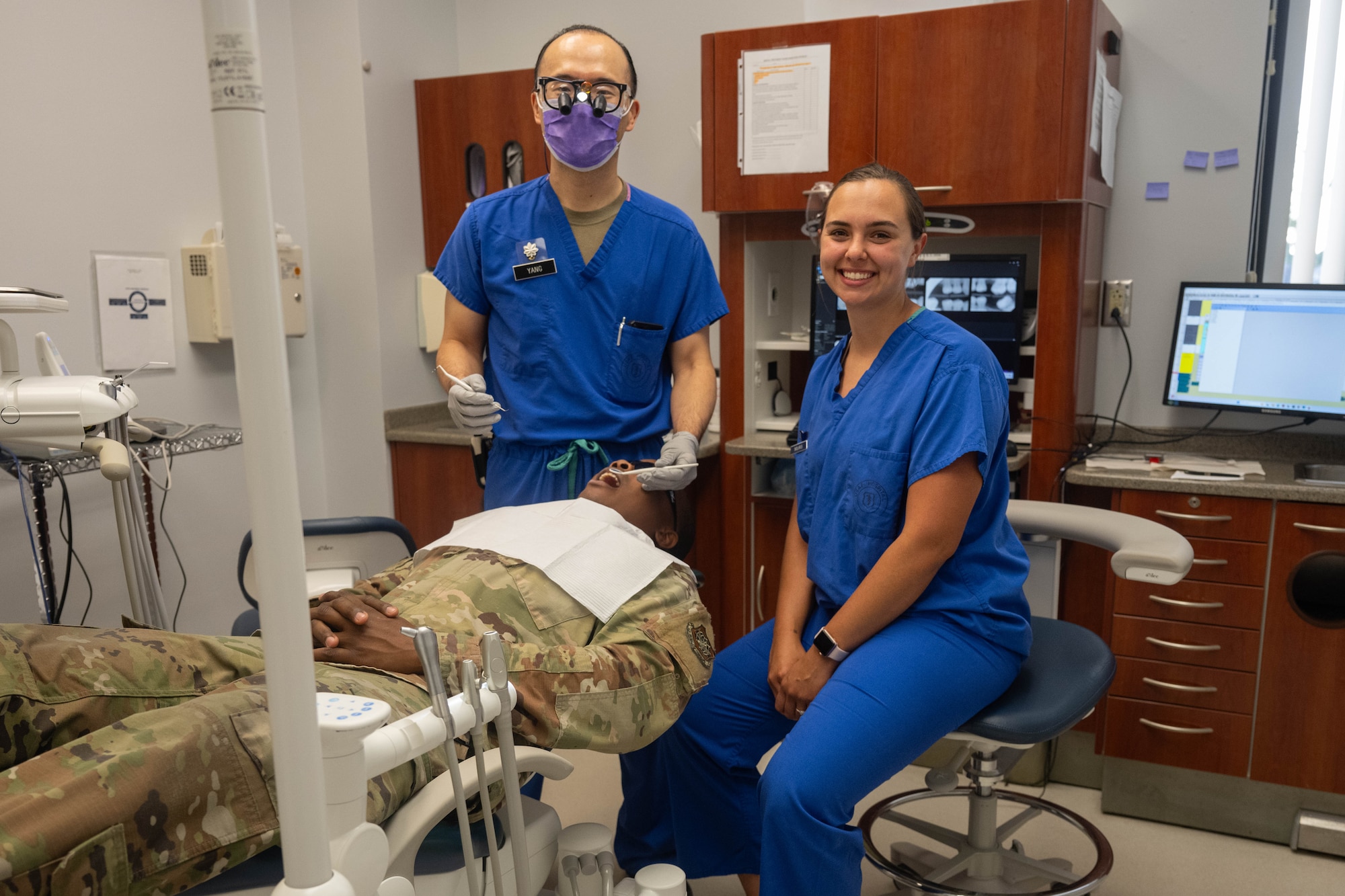 A man and a woman smile for a photo while providing dental care to a patient in uniform.
