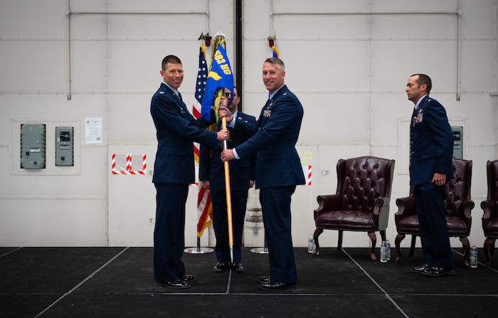 Two men in blue military service dress grip a squadron heraldry flag between them as they stand center stage and smile toward the camera.