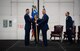 Two men in blue military service dress grip a squadron heraldry flag between them as they stand center stage and smile toward the camera.