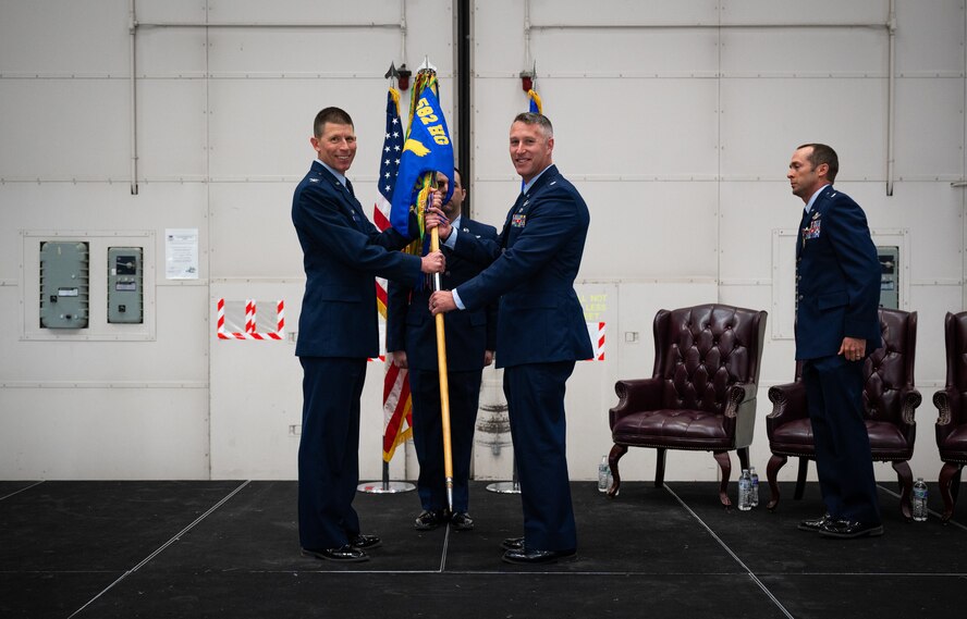 Two men in blue military service dress grip a squadron heraldry flag between them as they stand center stage and smile toward the camera.