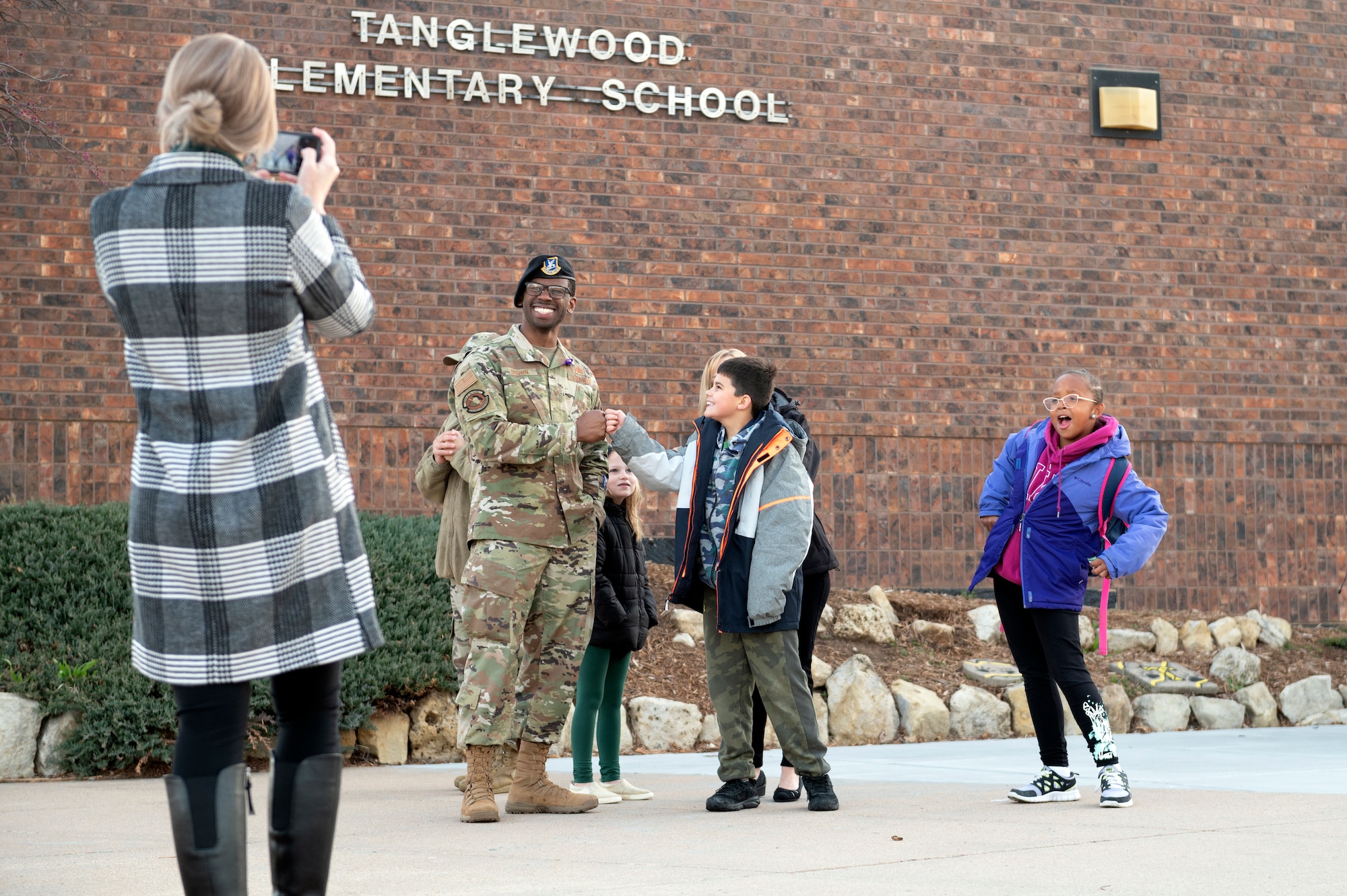 Tech. Sgt. Nicholas Brown, 22nd Security Forces Squadron, greets children at Tanglewood Elementary School on April 1, 2022, as a parent takes a photo and classmates pose for the camera. Several Team McConnell Airmen went to the school as part of a "Fist Bumps and High 5s' event, to welcome the arriving students with smiles, fist bumps and high fives to brighten their day. (Air Force Photo by Master Sgt. John Gordinier)