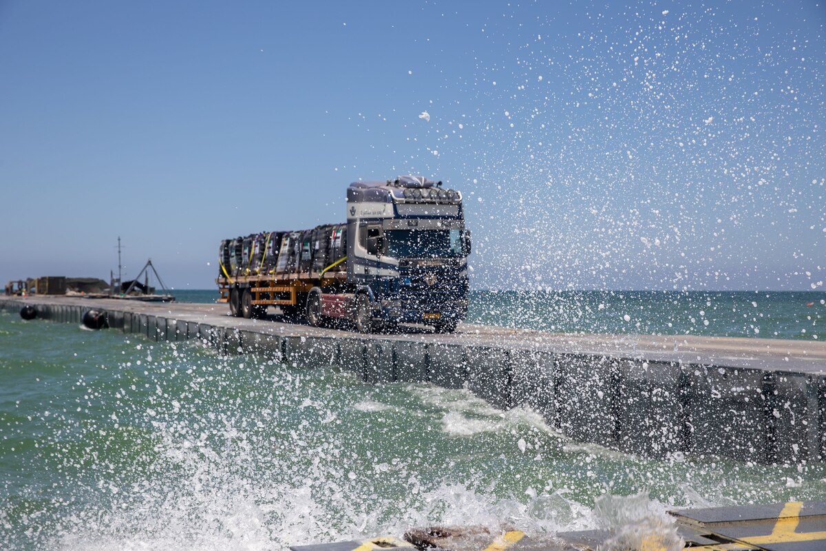 A truck carrying cargo drives across a pier toward a shoreline structure where seawater splashes.