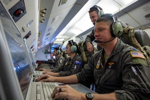 Four people sit at the on-board mission console of an aircraft while another man stands behind them.