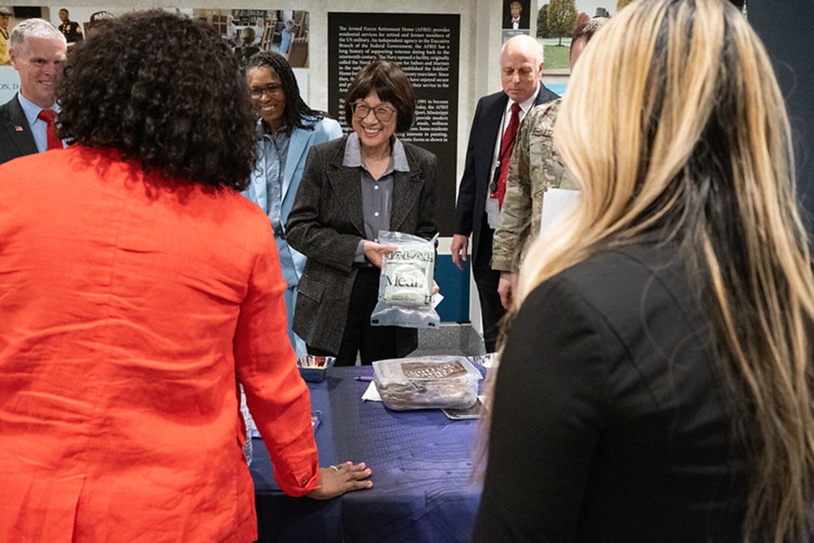 A person stands behind a table, holding a bag of food as people watch.