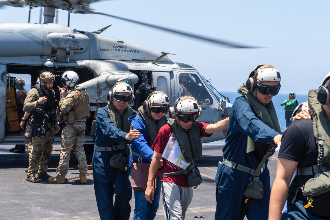 Sailors wearing flight helmets guide civilians in helmets away from a helicopter parked on a flight deck.