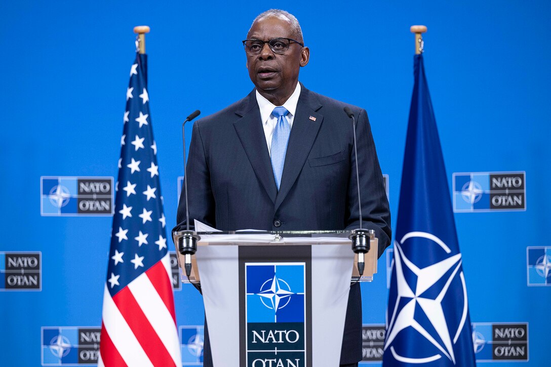 Secretary of Defense Lloyd J. Austin III speaks at a lectern, with a U.S. and a NATO flag behind him.