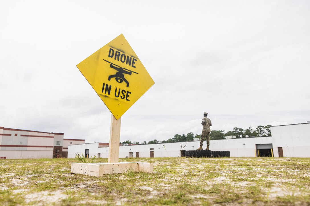 A yellow diamond-shaped sign reading "DRONE IN USE" stands in a field where a Marine works in the distance.
