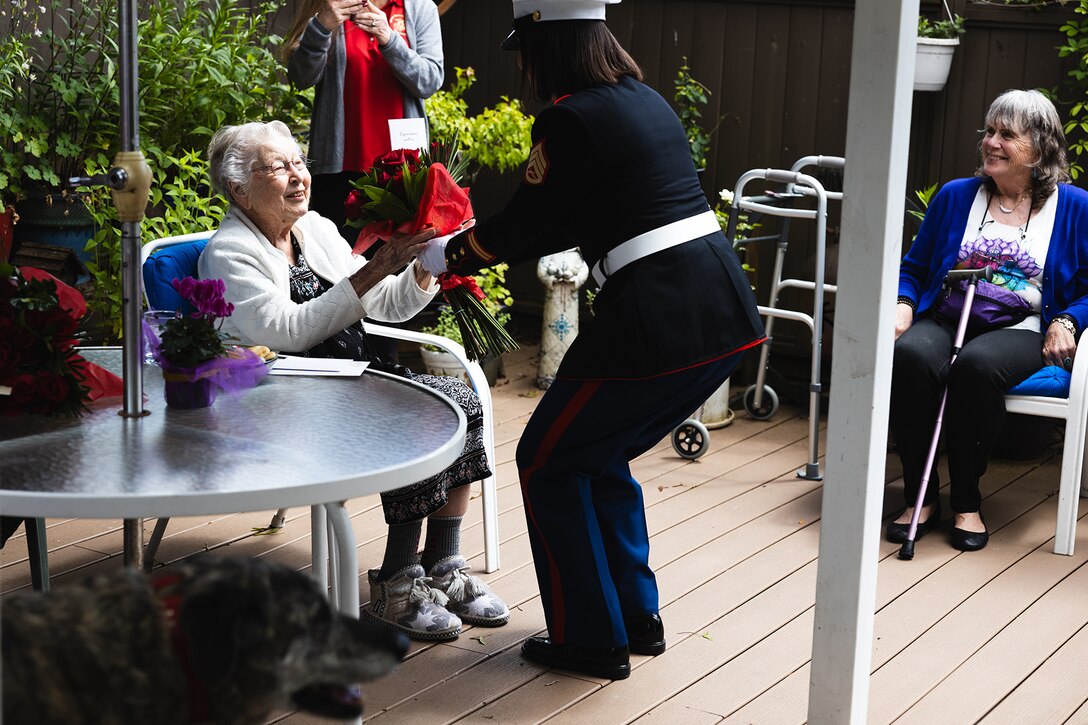 A Marine presents red roses to a seated World War II veteran as other people and a dog watch.