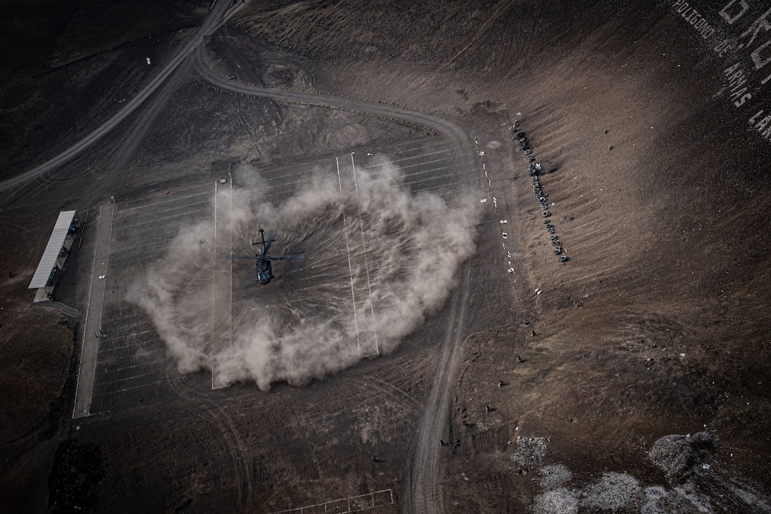 A helicopter takes off from a desert landscape, kicking up a plume of dust.