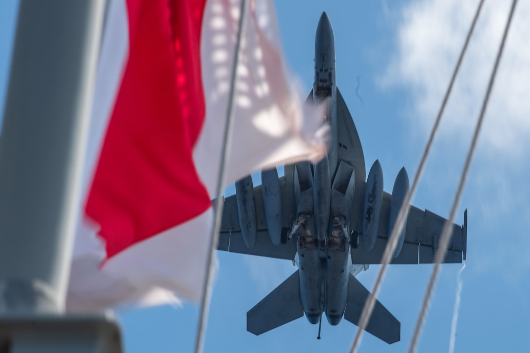 An aircraft flies over ship as seen in between a flying flag and ropes on the ship.