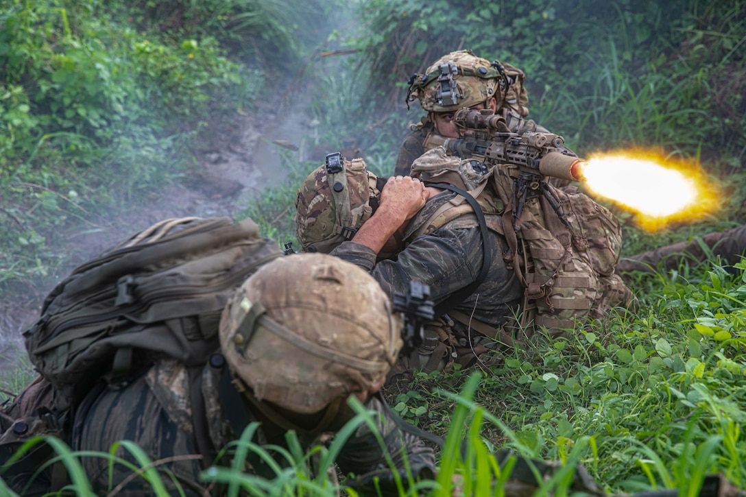 Two soldiers take cover in a jungle as a fellow service member fires a gun, creating a fireball.