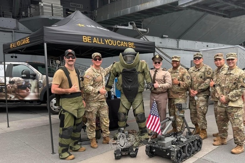 Service members pose with robotic devices under a tent that reads "Be All You Can Be."