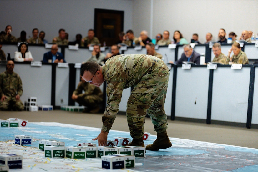 A soldier bends to place a small box on a large map during a training as other people watch.