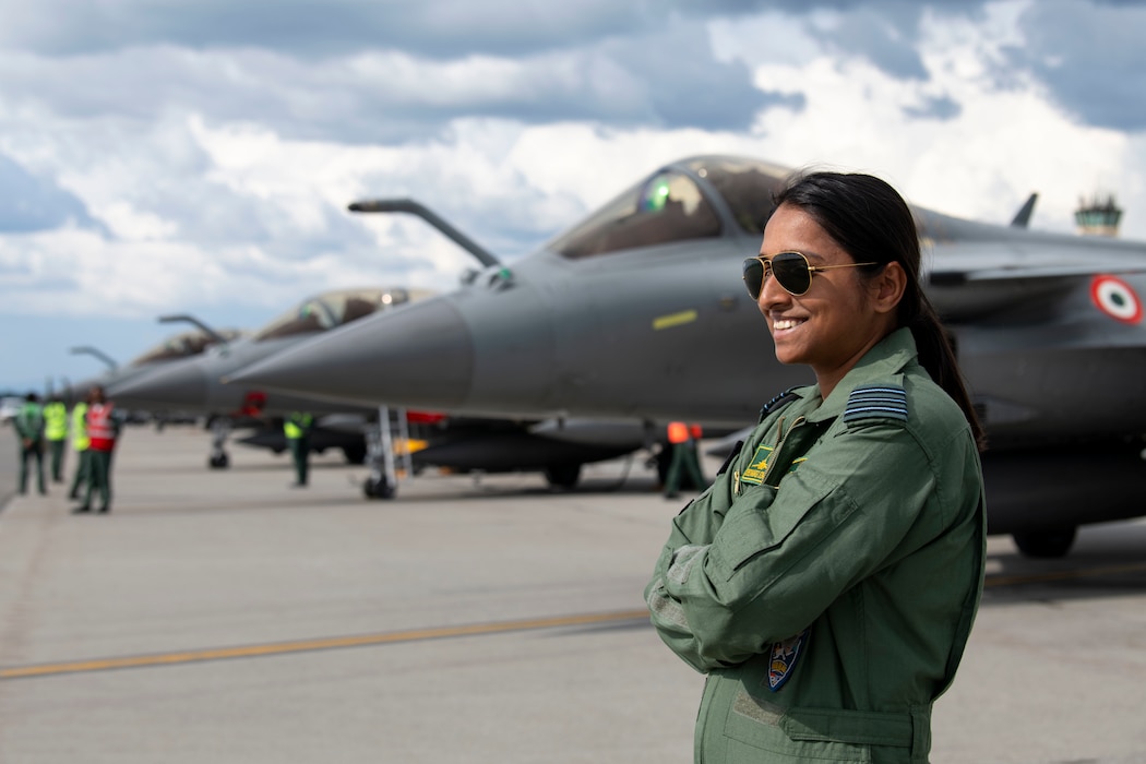 A female pilot from the Indian Air Force poses for a photo in front of 4 Rafale aircraft lined up diagonally behind her.