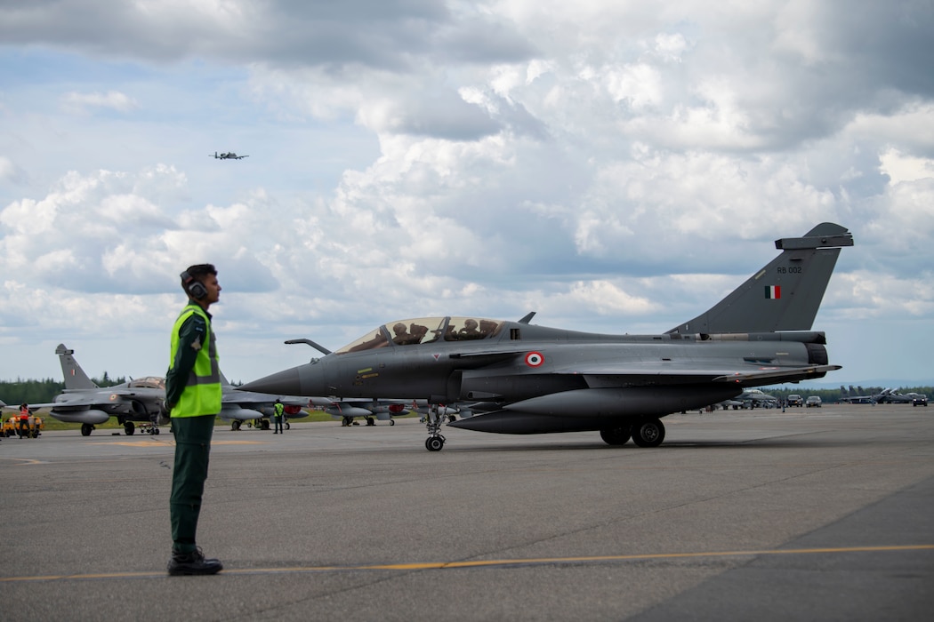A maintainer from the Indian Air Force crosses his hands behind his back in front of a Rafale Aircraft taxiing. An A-10 aircraft flies in the sky in the background.