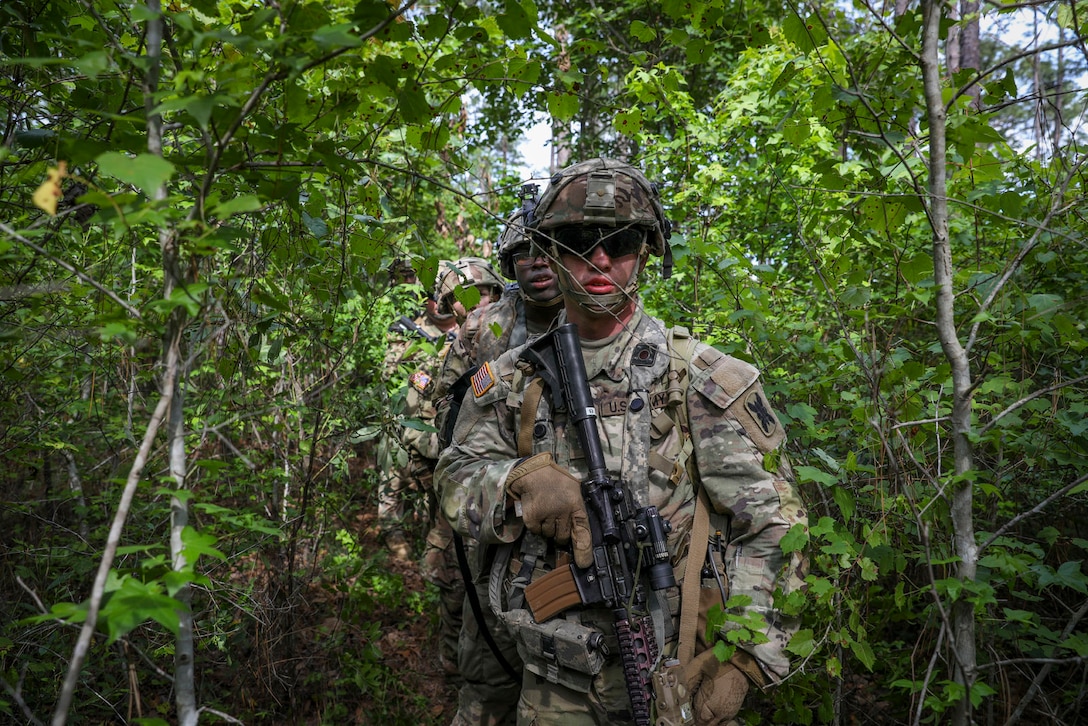 Soldiers holding weapons look forward while standing in a loose line in a forested area.