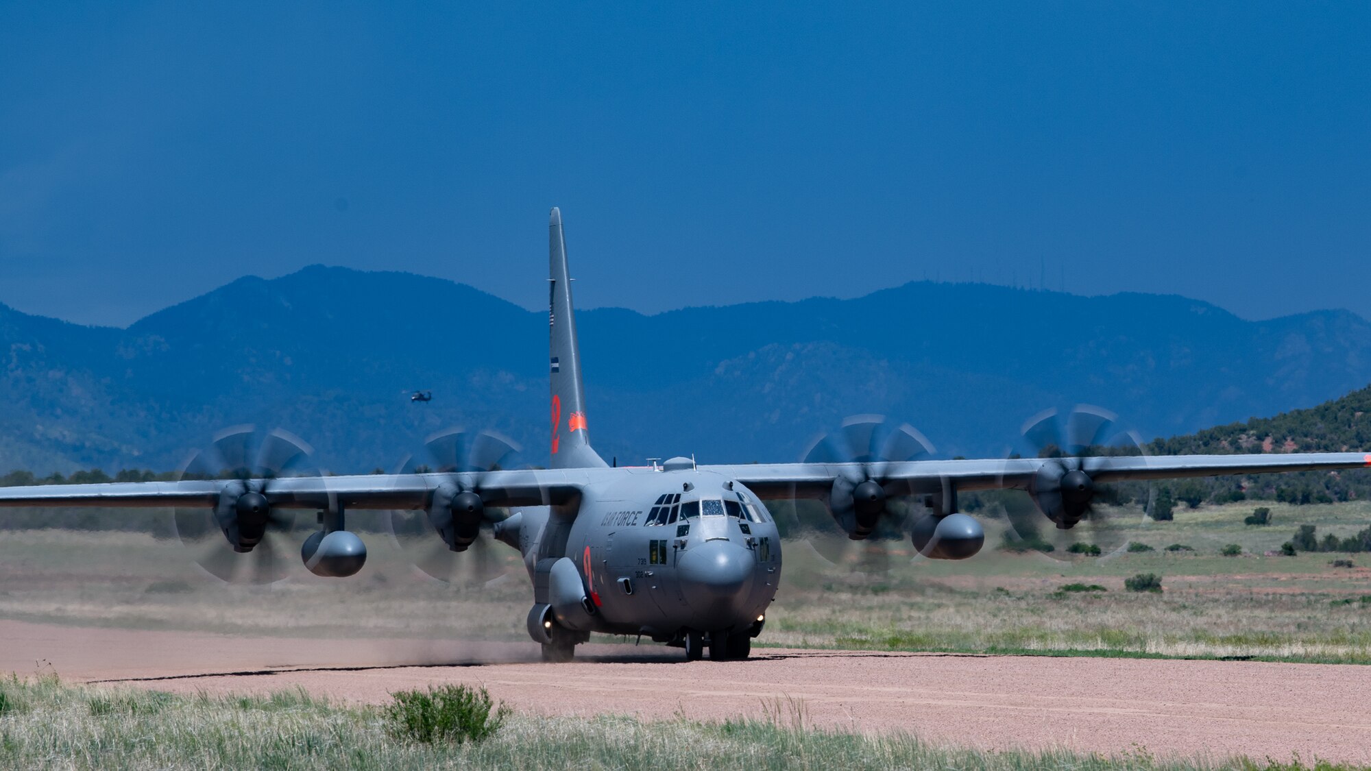 A military cargo aircraft takes off at a dirt airstrip with a second aircraft in the background.
