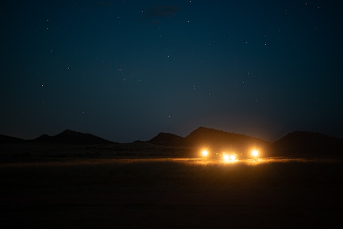 A military aircraft with bright lights on a dirt landing strip against a mountainous starry background at night.