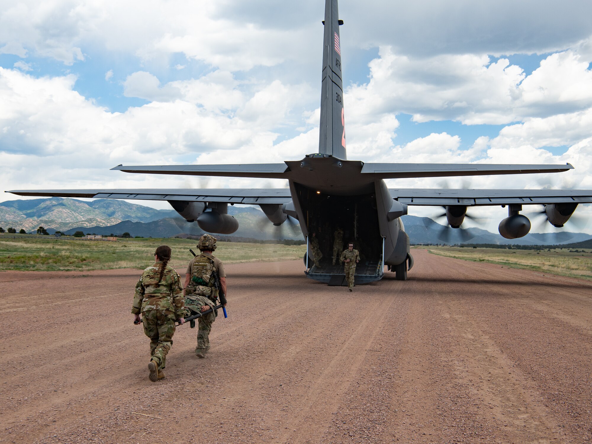 Two servicemembers with a medical litter walking towards the open cargo bay of a military aircraft with its engines running on a dirt airstrip.