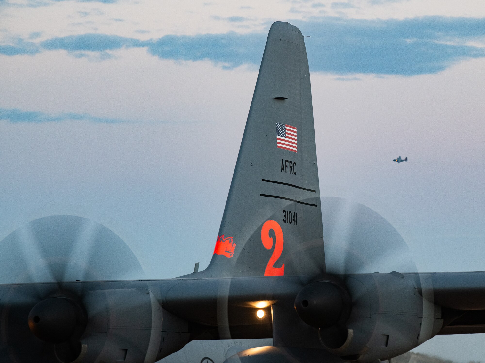 The tail of a military cargo aircraft with spinning propellers in the foreground and a second aircraft flying in the background.