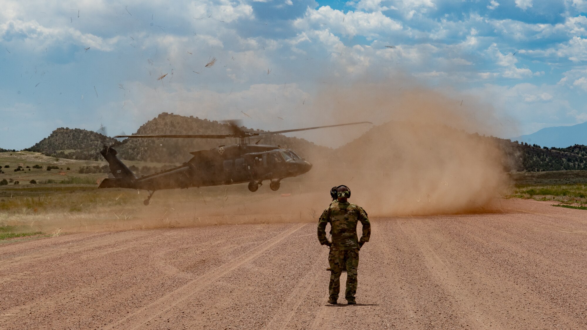 A soldier stands on a dirt landing strip watching a helicopter kicking up dust as it lands.