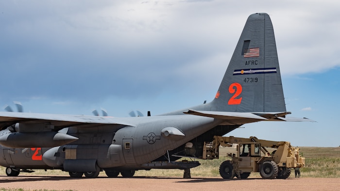 A forklift loading a piece of equipment into the cargo bay of a military aircraft with its engines running on a dirt airstrip.