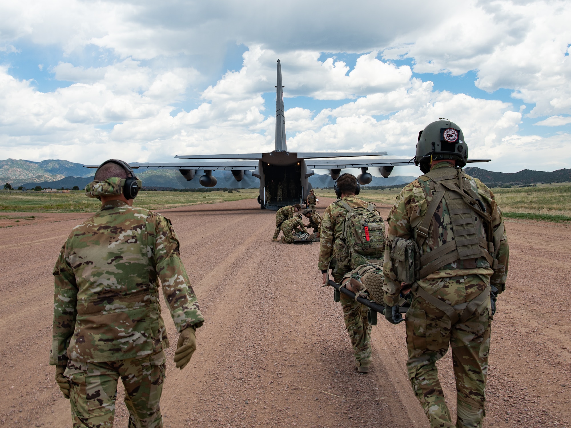 Servicemembers walk towards a military aircraft with its engines running while carrying a litter on a dirt airstrip.