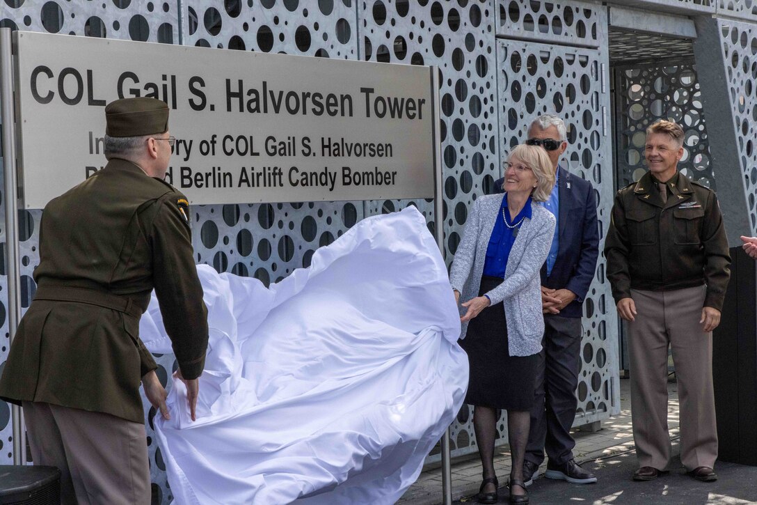 A military officer and a civilian remove a sheet of white plastic from a large metal sign in front of a tower.