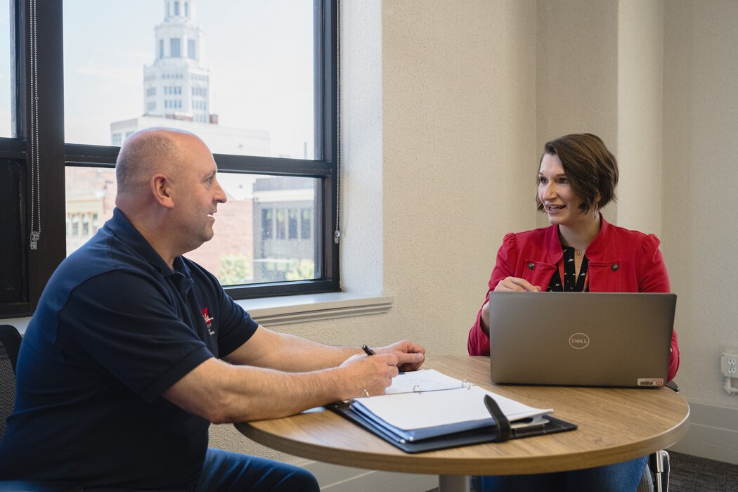 A man and woman sit on opposite sides of a table, with a notebook and laptop between them.