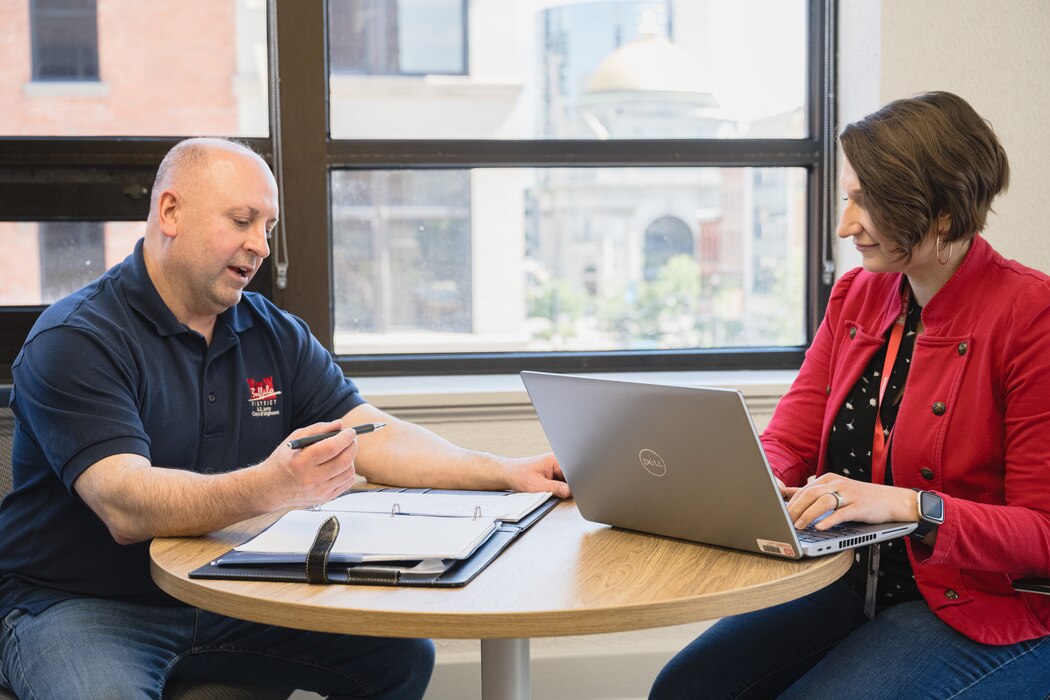 A man and woman sit on opposite sides of a table, with a notebook and laptop between them.