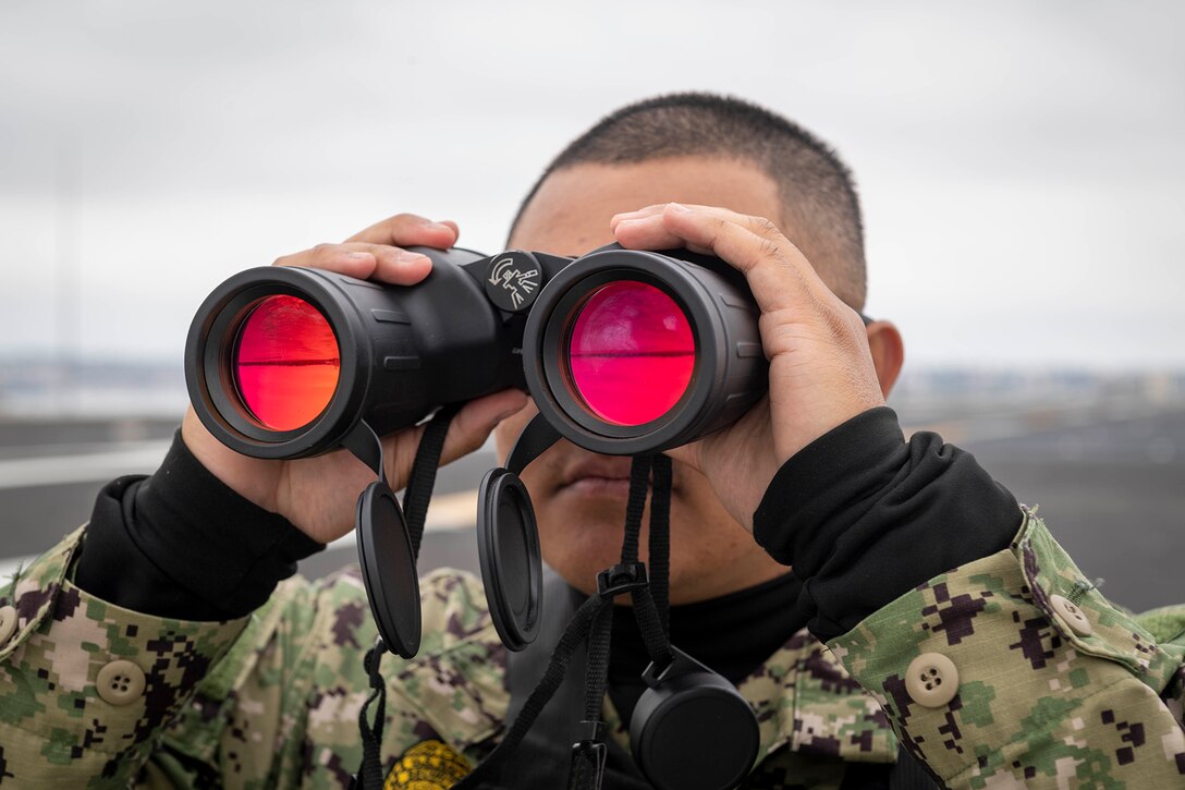 Close-up of a sailor looking through large binoculars while standing on a ship’s flight deck.