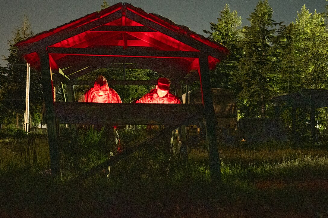 Soldiers look down at materials under a gazebo in a wooded area at night. The are illuminated by a red light.