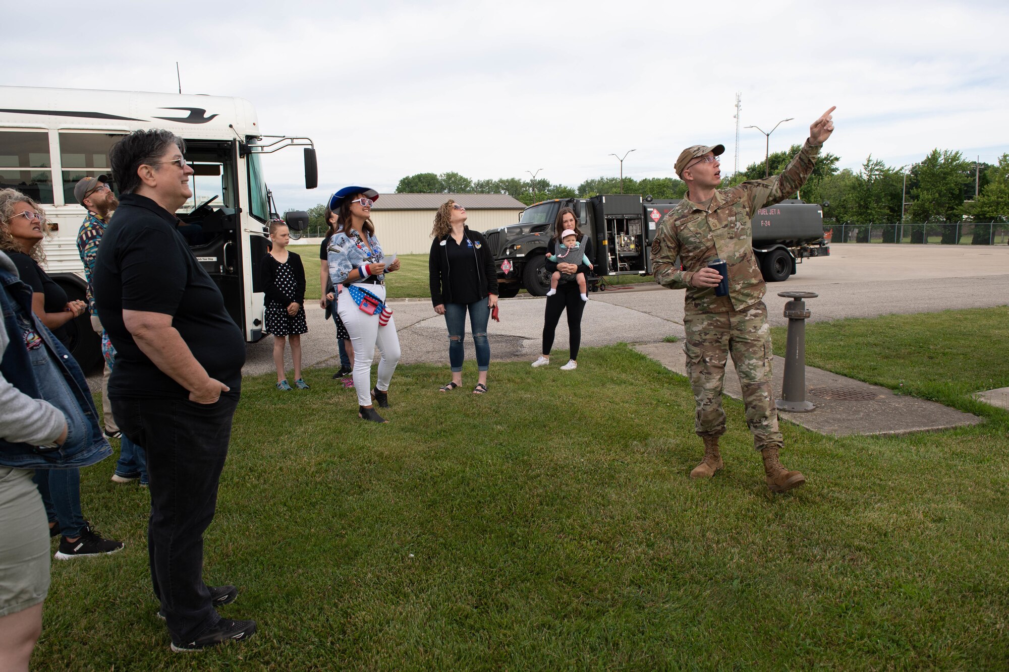 A man in a military uniform points upwards, with a crowd of civilians behind him looking in the direction of his hand. In the background, a white bus and a military fuels vehicle.
