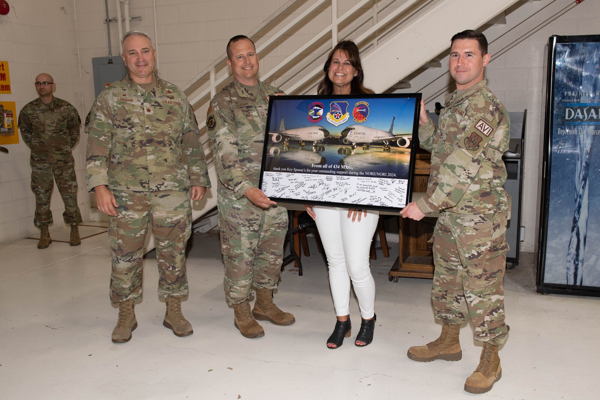 A woman dressed in civilian attire holds a frame and poses for a photo with three men dressed in military uniforms. In the far background, a man also in uniform.
