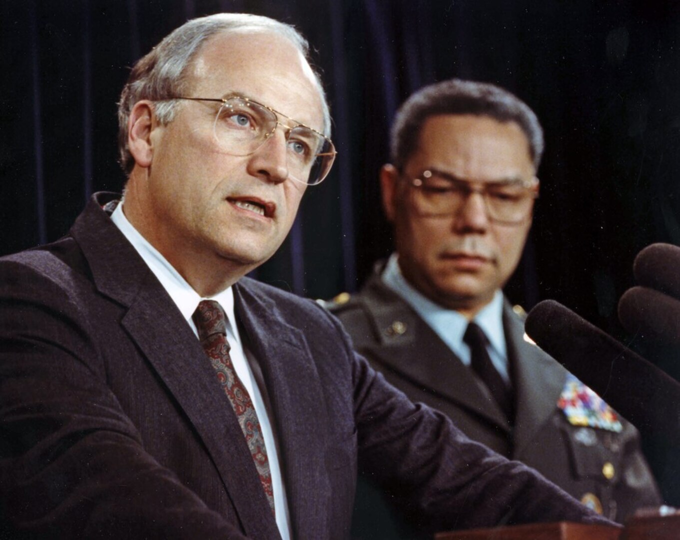 White man in foreground speaks into a microphone with a black man standing behind him