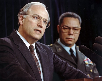 White man in foreground speaks into a microphone with a black man standing behind him