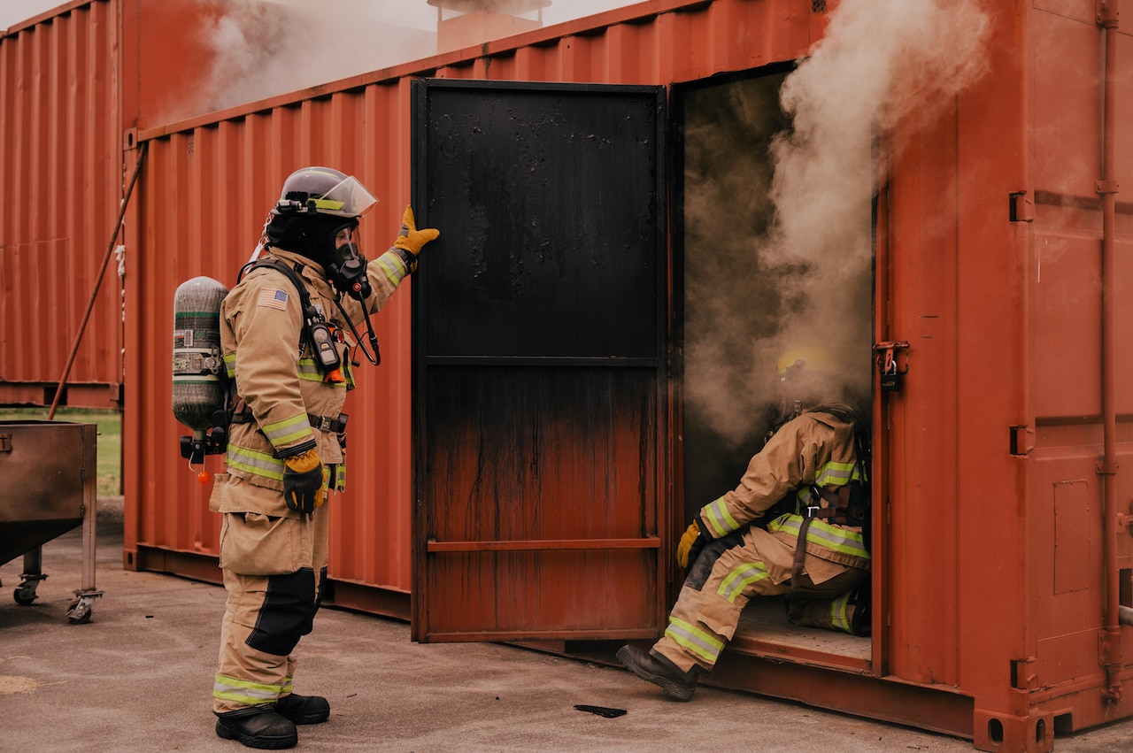A firefighter leans into a smoke-filled container, as another firefighter watches.