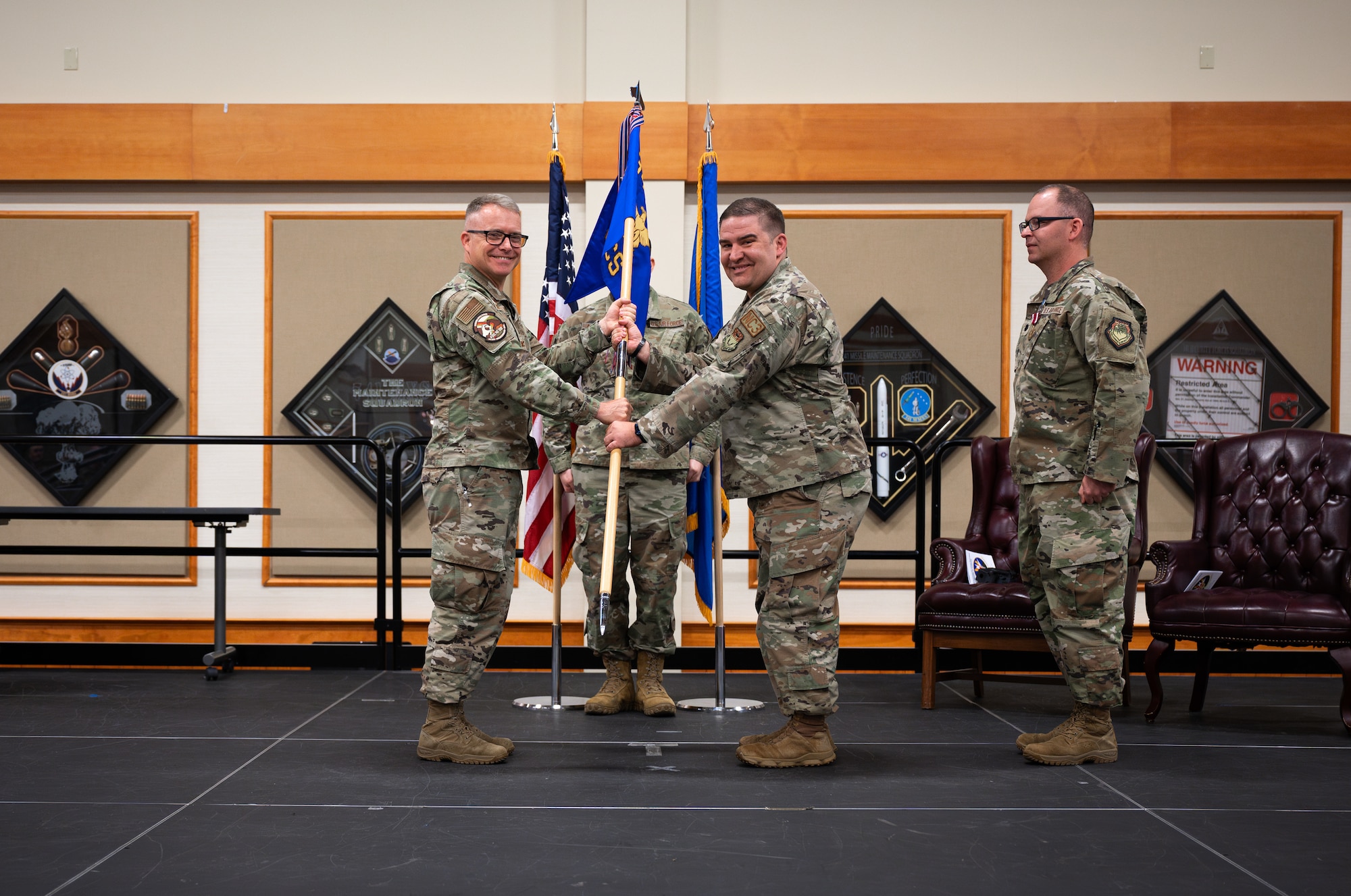 Two men in military uniform stand centerstage as they grip a guidon flag between them and look toward the camera.