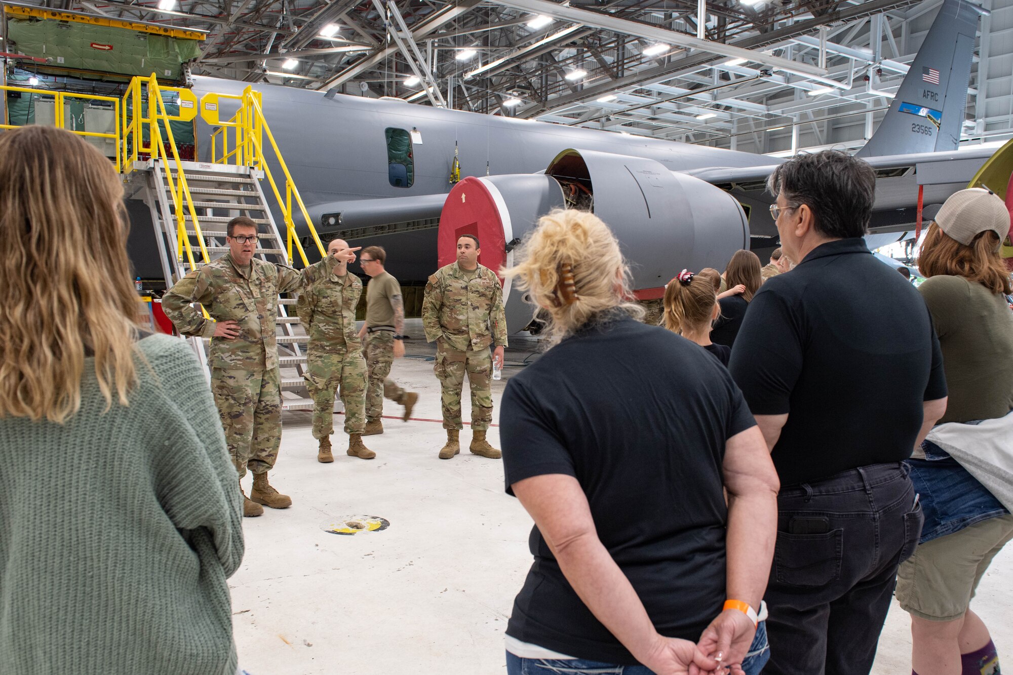 People in civilian clothing listen to a man in a military uniform speak. In the background, a military plane with portable stairs attached to the side of the aircraft.
