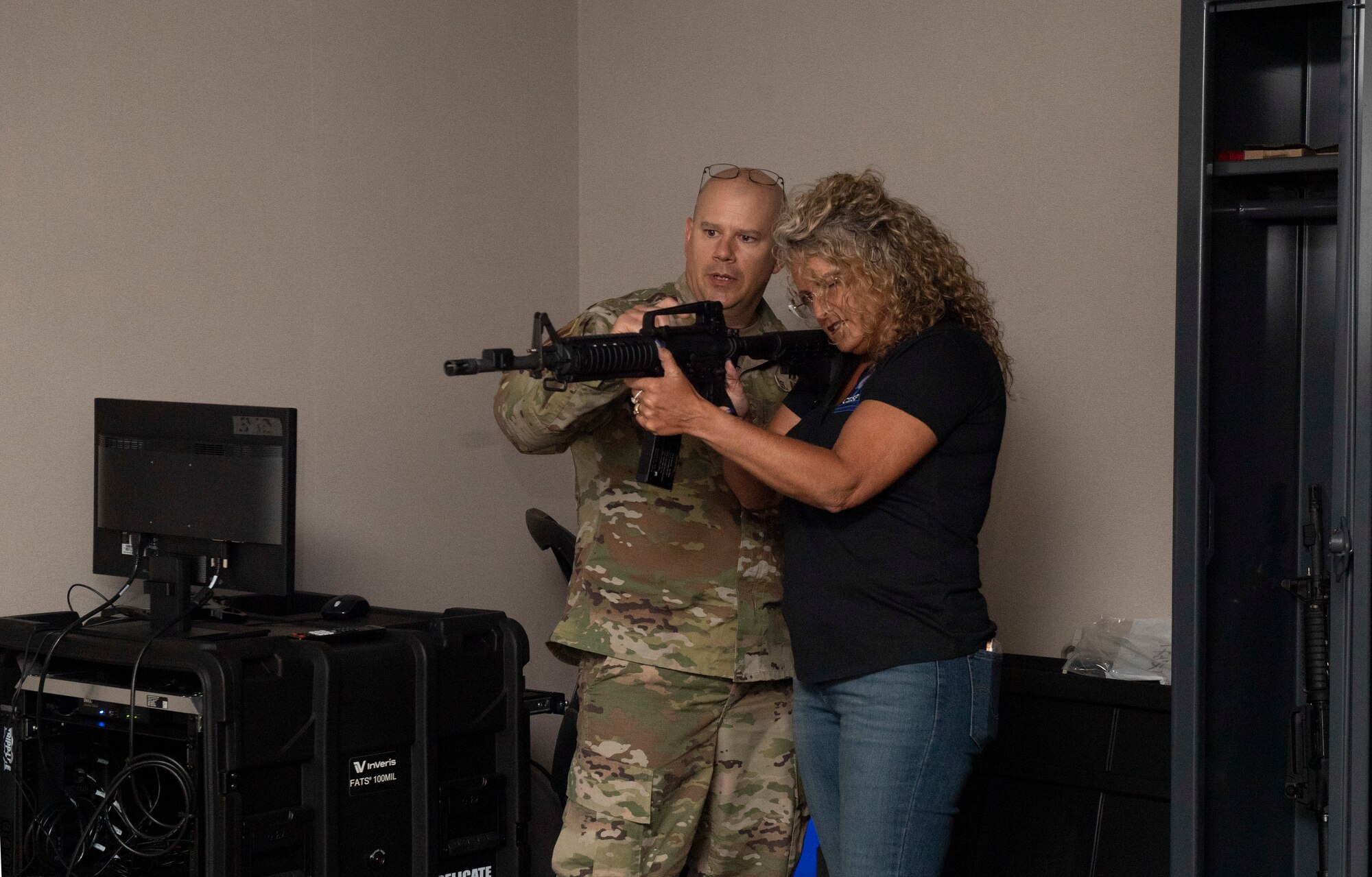 A woman in civilian attire holds a mock military weapon as a man in a military uniform instructs her on usage and components.
