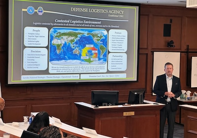 A man stands at the front of a room with a screen projector behind him showing a map of the world