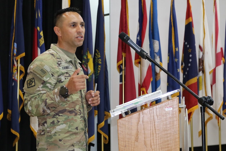 A male U.S. Soldier gestures naturally behind a light wood podium with a black microphone.