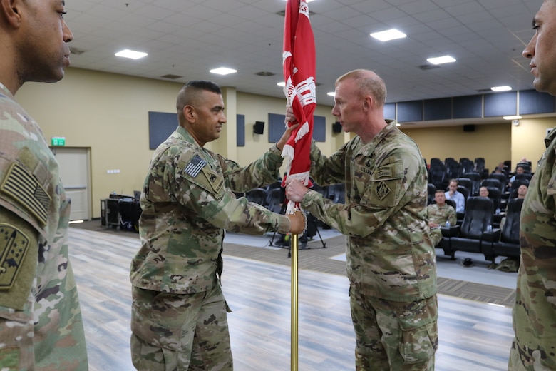 Two uniformed US. Army Soldiers stand facing each other. Together they are holding a gold flagpole vertically between them. The flagpole has a red flag with white fring wrapped around it.