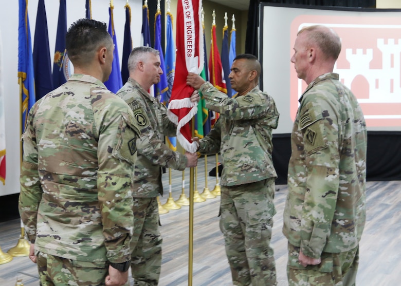 Four male U.S. soldiers in uniform standing in a circle, with one soldier passing a long gold flagpole wrapped with a red with white fringe to another soldier.