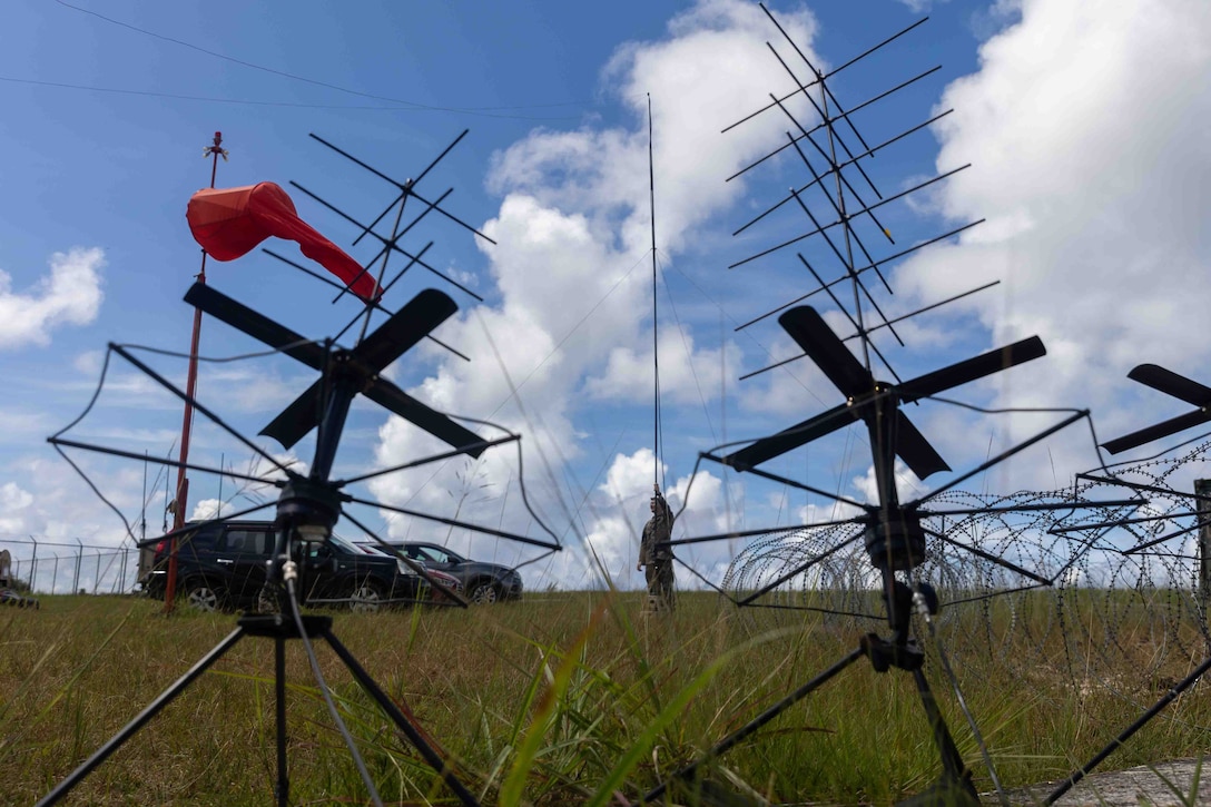 A Marine stands in a large field holding a pole surrounded by small satellites and some parked cars.