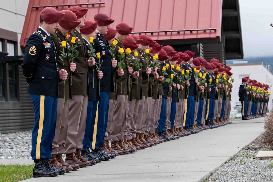 A long line of soldiers wearing red berets stand with their heads bowed holding yellow roses.